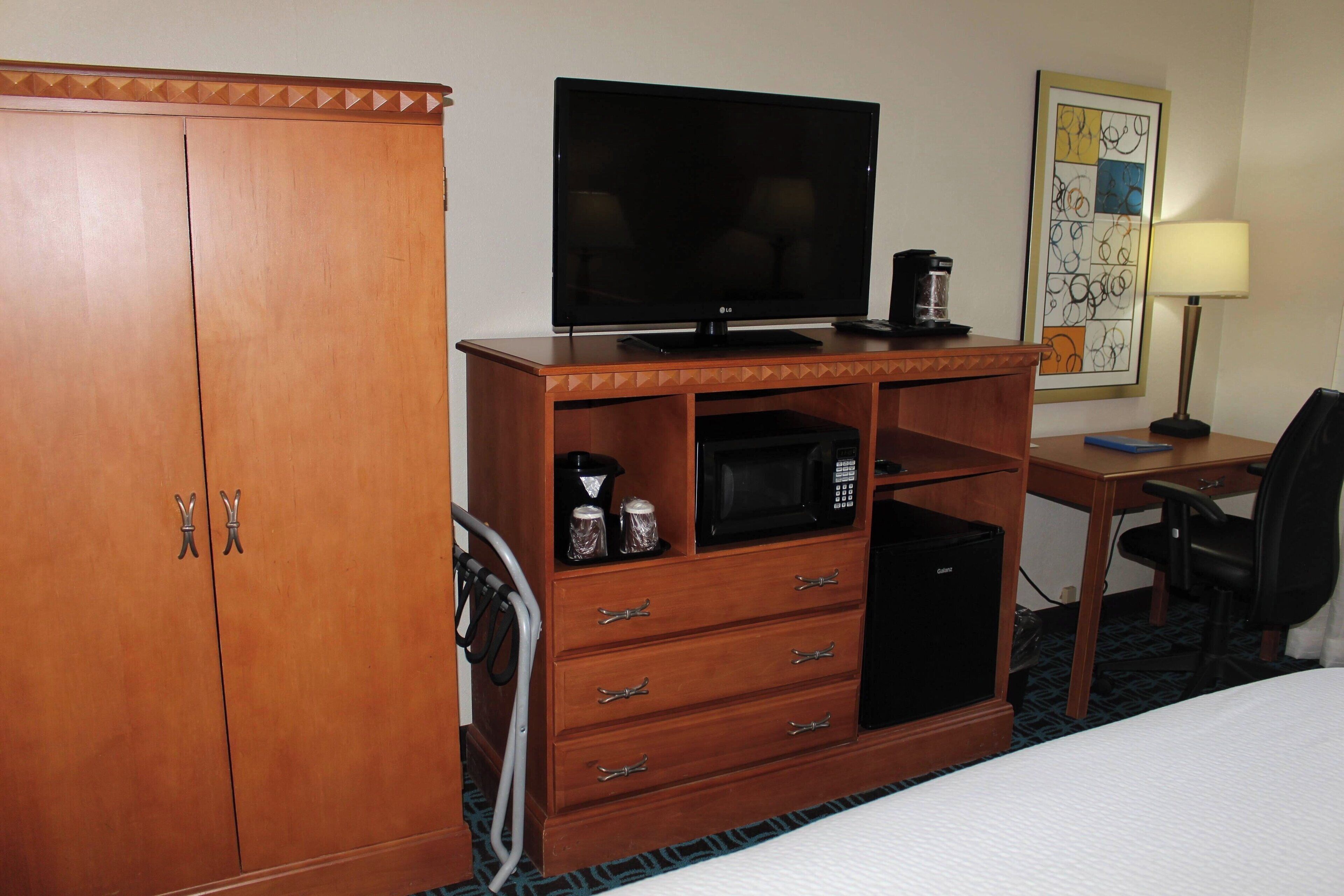 Una habitación de hotel con armario de madera, televisor de pantalla plana, cafetera, microondas, escritorio con silla y cama.