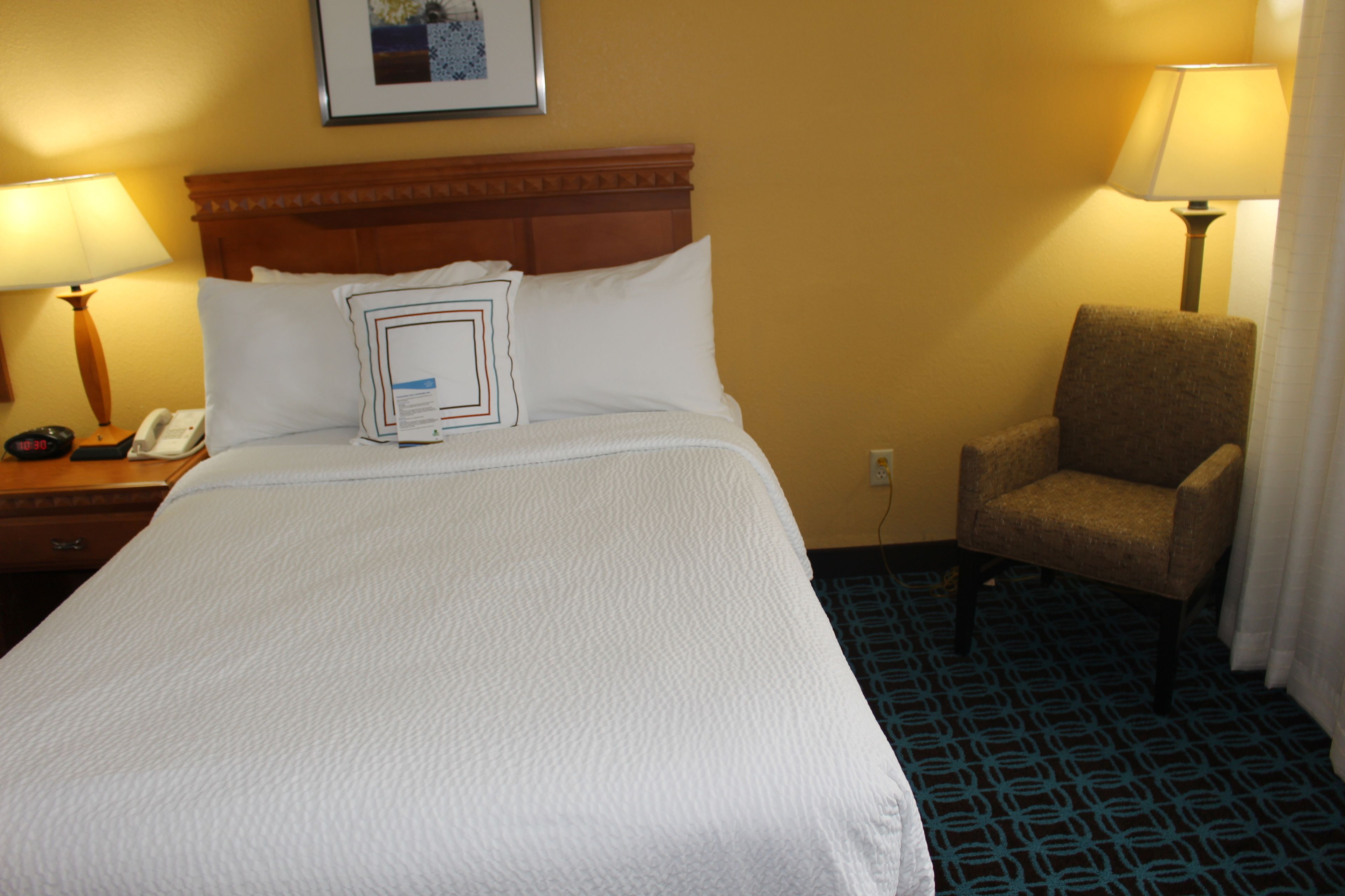 Habitación de hotel con una cama grande, cabecera de madera, dos lámparas de noche, una silla y alfombra estampada.