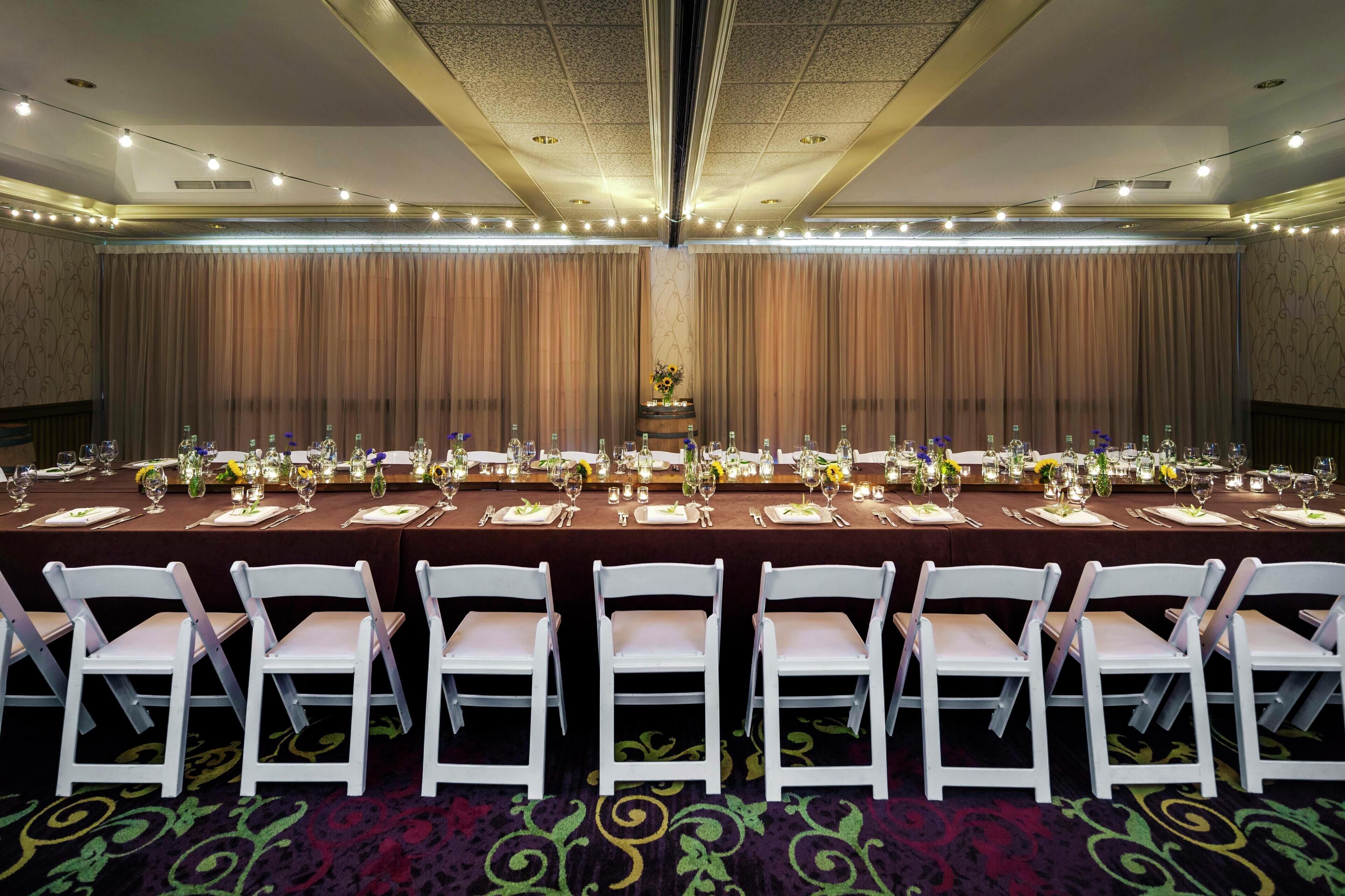 Un salón de conferencias con una larga mesa dispuesta para una reunión, rodeada de sillas blancas, cortinas y un centro de mesa con flores.