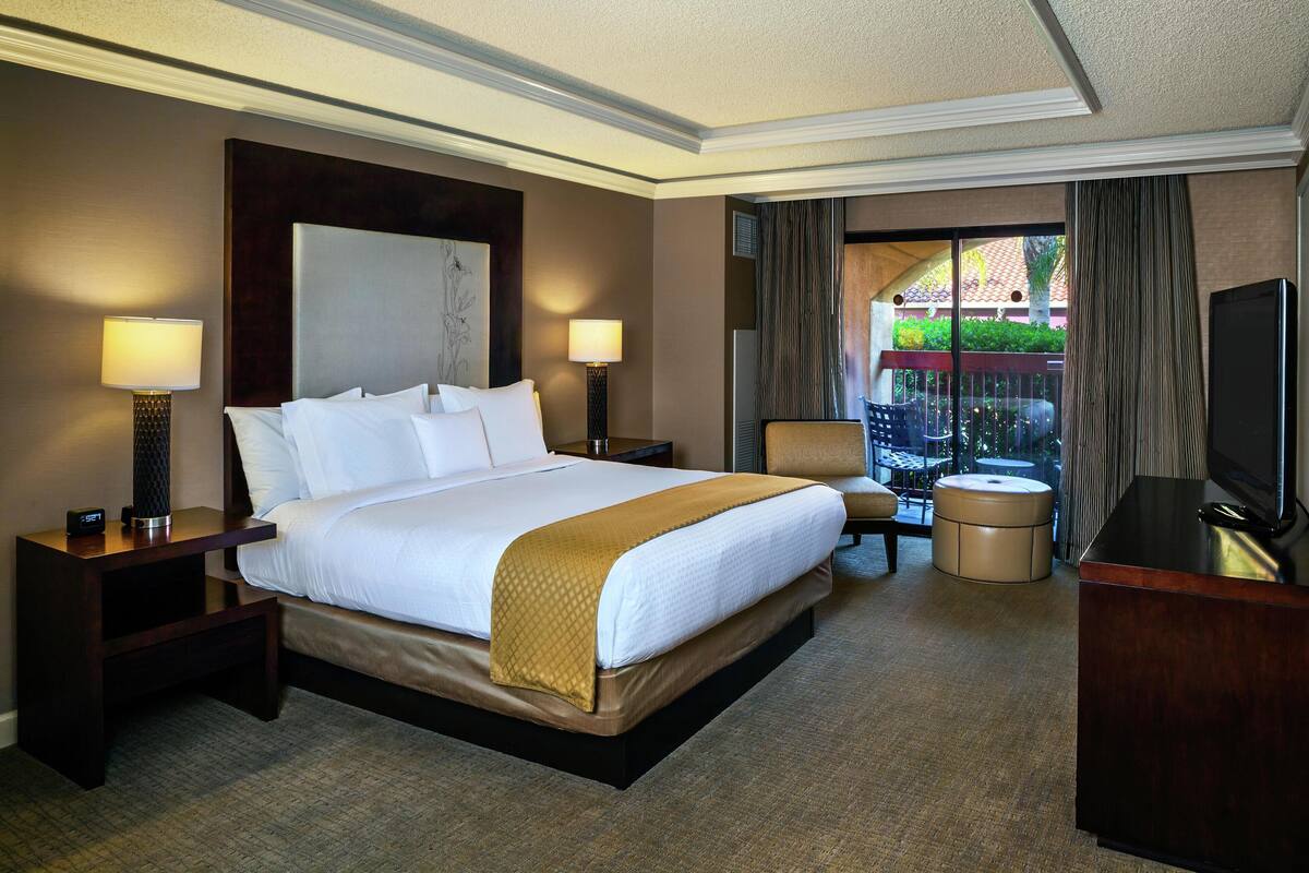 Une chambre d’hôtel dotée d’un grand lit, de deux tables de chevet avec des lampes, d’une télévision, d’un coin salon avec un fauteuil et un pouf, et offrant une vue sur l’extérieur par une fenêtre.