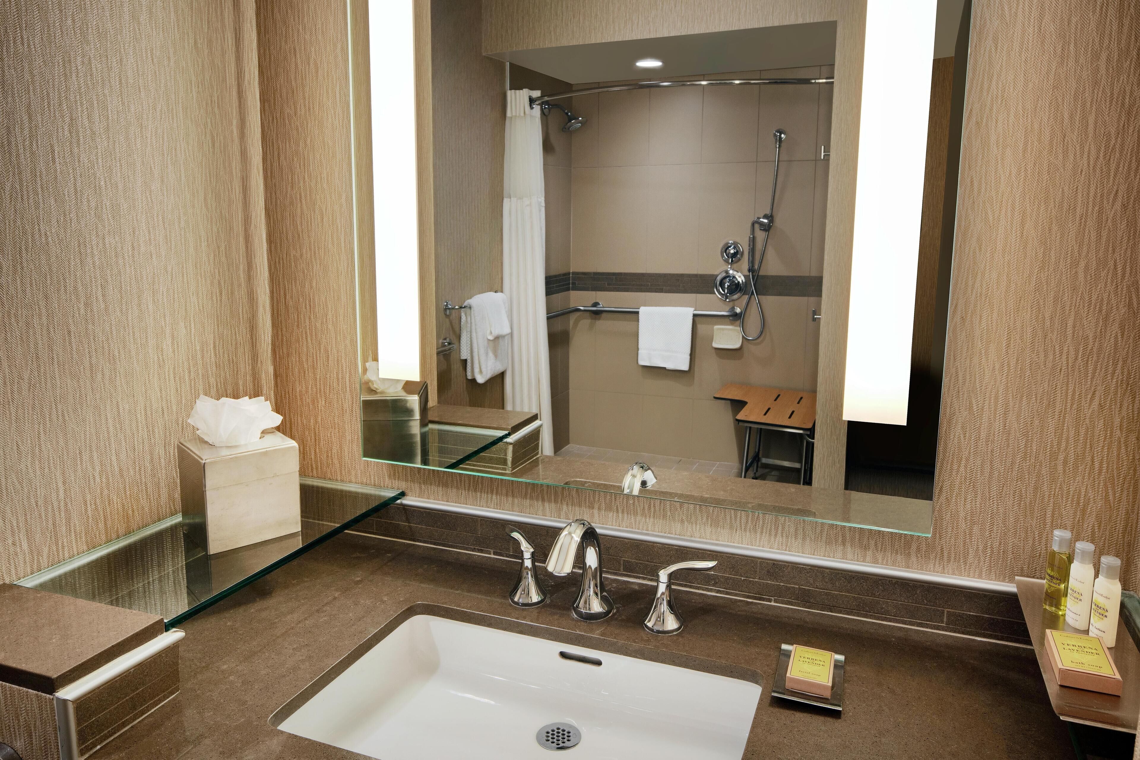 A modern bathroom with a large mirror, a glass shower enclosure, a sink with two faucets, and toiletries on the counter.