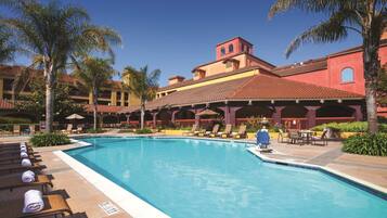 A pool area with lounge chairs and umbrellas, a clear blue sky, and a building with red-tiled roofs and balconies.