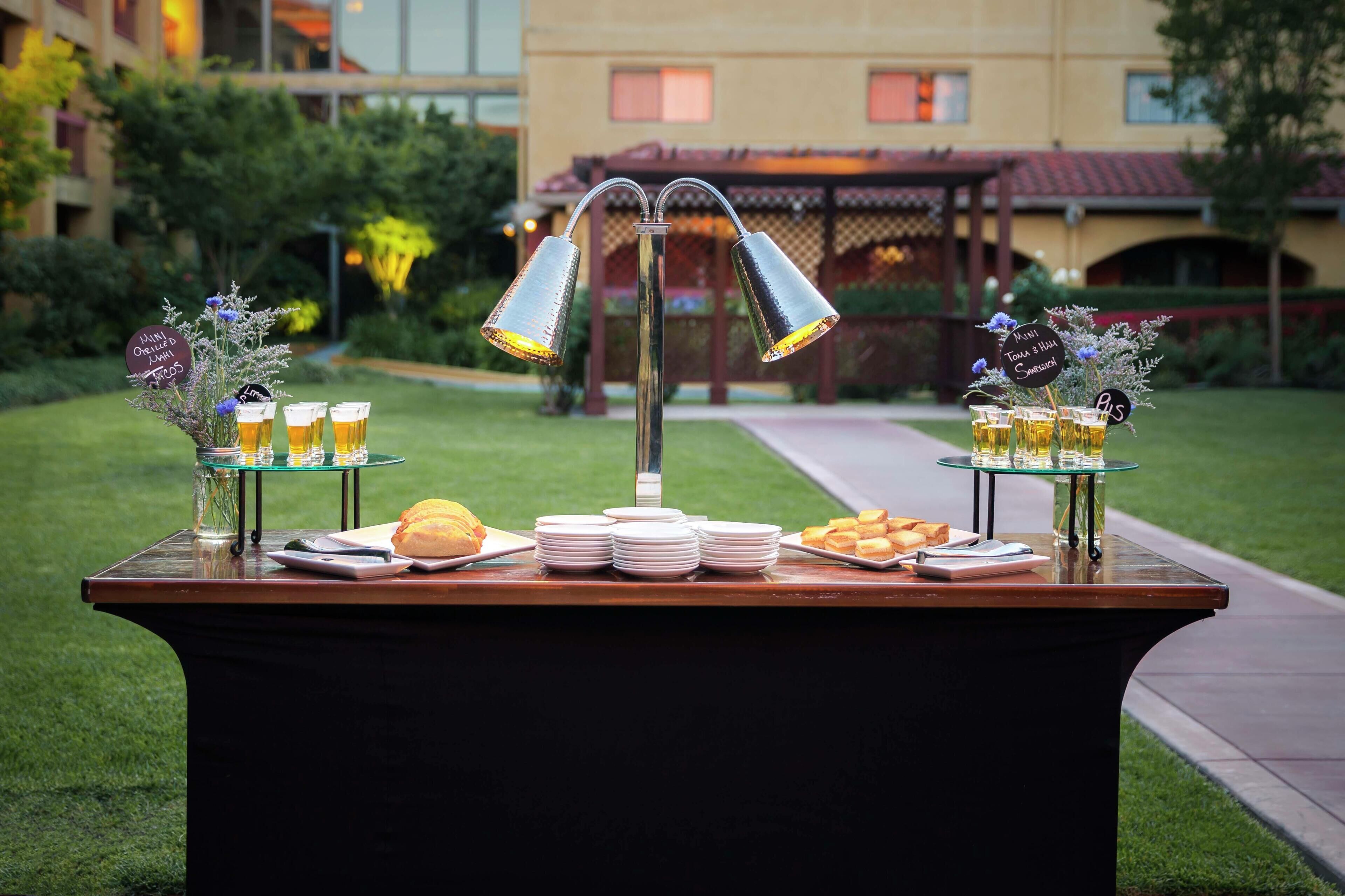 Mesa de buffet al aire libre con comida y bebidas, rodeada de follaje y un edificio al fondo.