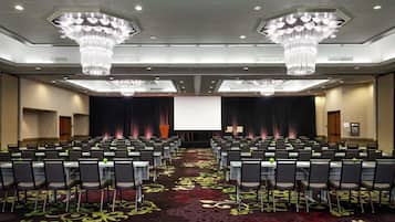 Un salón de conferencias con filas de mesas y sillas, una pantalla grande al frente y candelabros en el techo.
