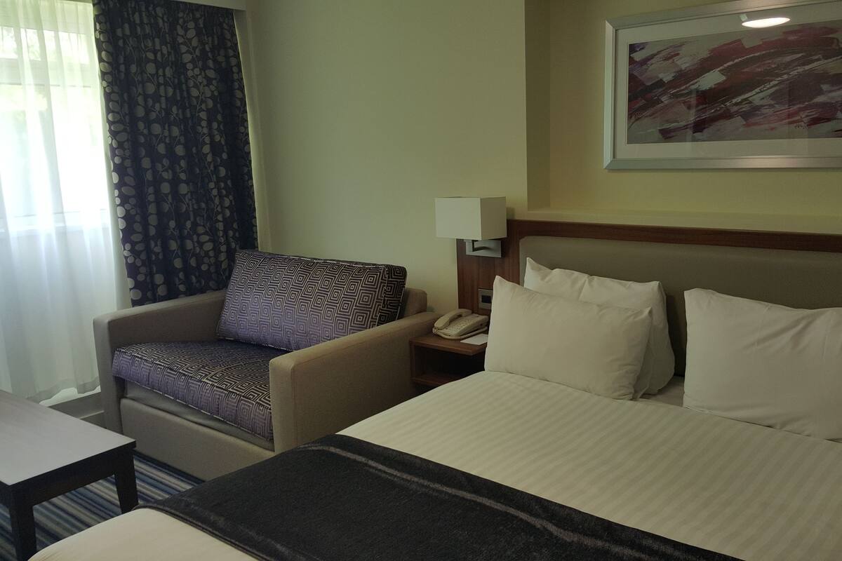 A hotel room with a bed, a chair, a nightstand, and a painting on the wall.