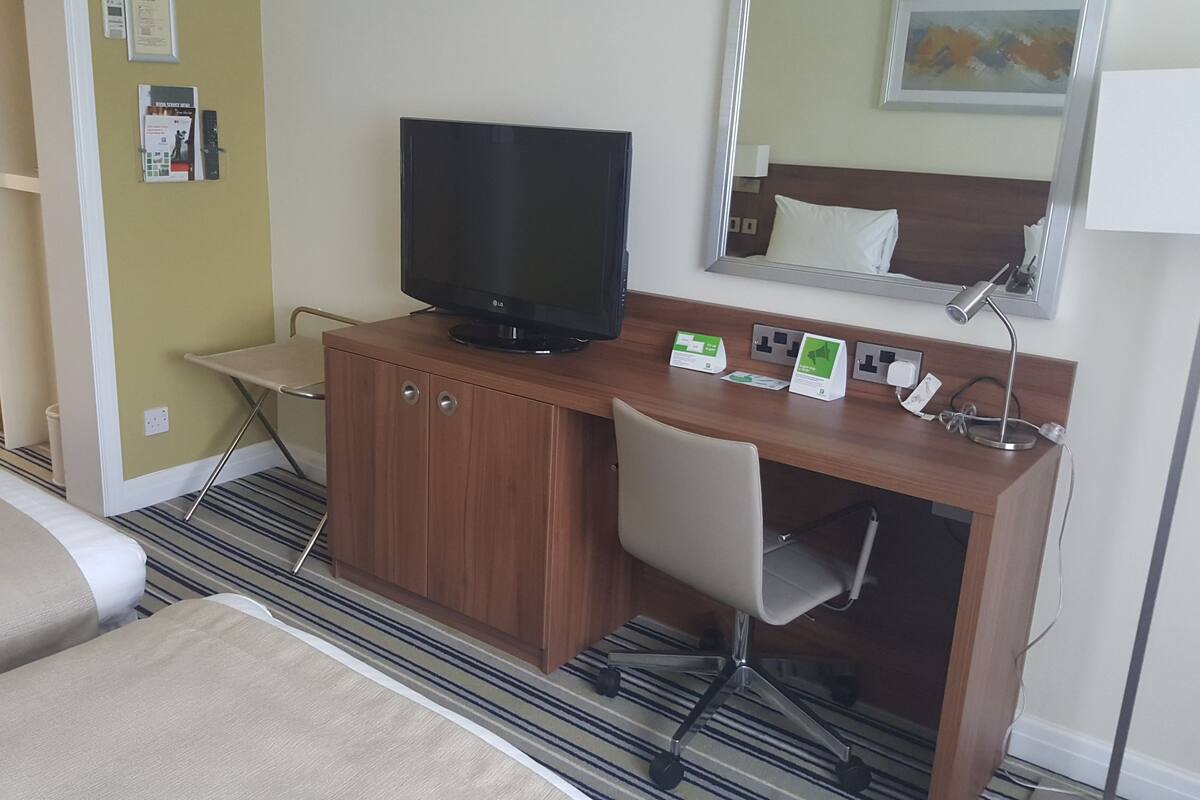 A hotel room with a bed, a desk with a television, a chair, a desk lamp, and a mirror.