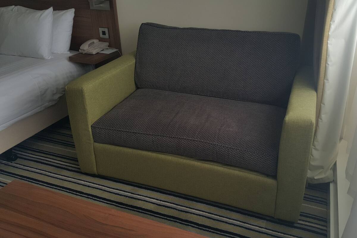 A hotel room with a bed, a small desk with a phone, and a grey armchair.