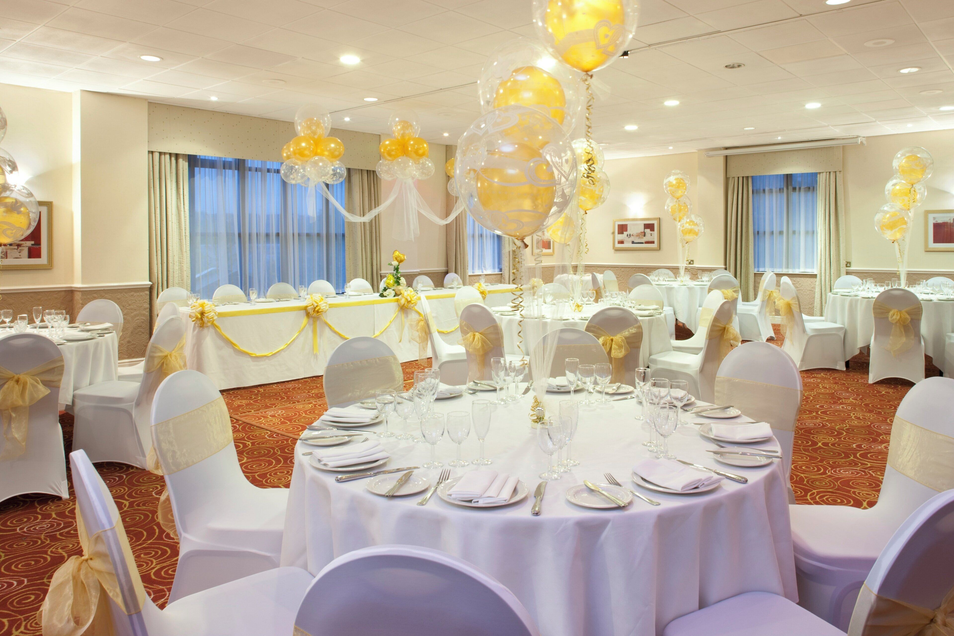 Un salón de banquetes con mesas redondas dispuestas para cenar, sillas blancas y cintas amarillas.
