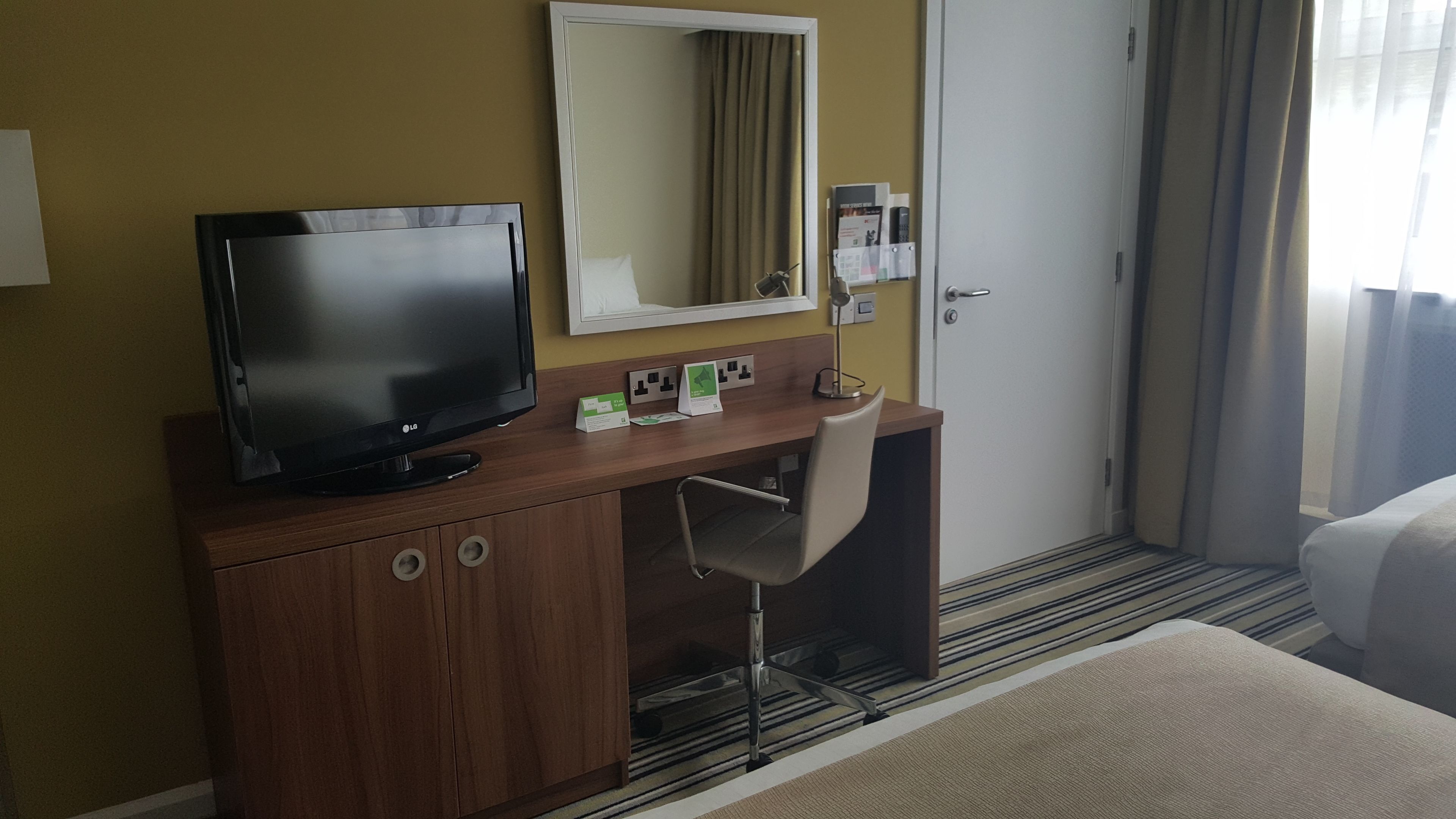 Una habitación de hotel con un escritorio de madera, televisor, espejo, silla y cama.