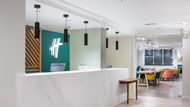 A modern reception area with a marble counter, pendant lighting, and a view into a dining area with colorful chairs and tables. - Holiday Inn Derby Nottingham M1 J25 by IHG (Nottingham)