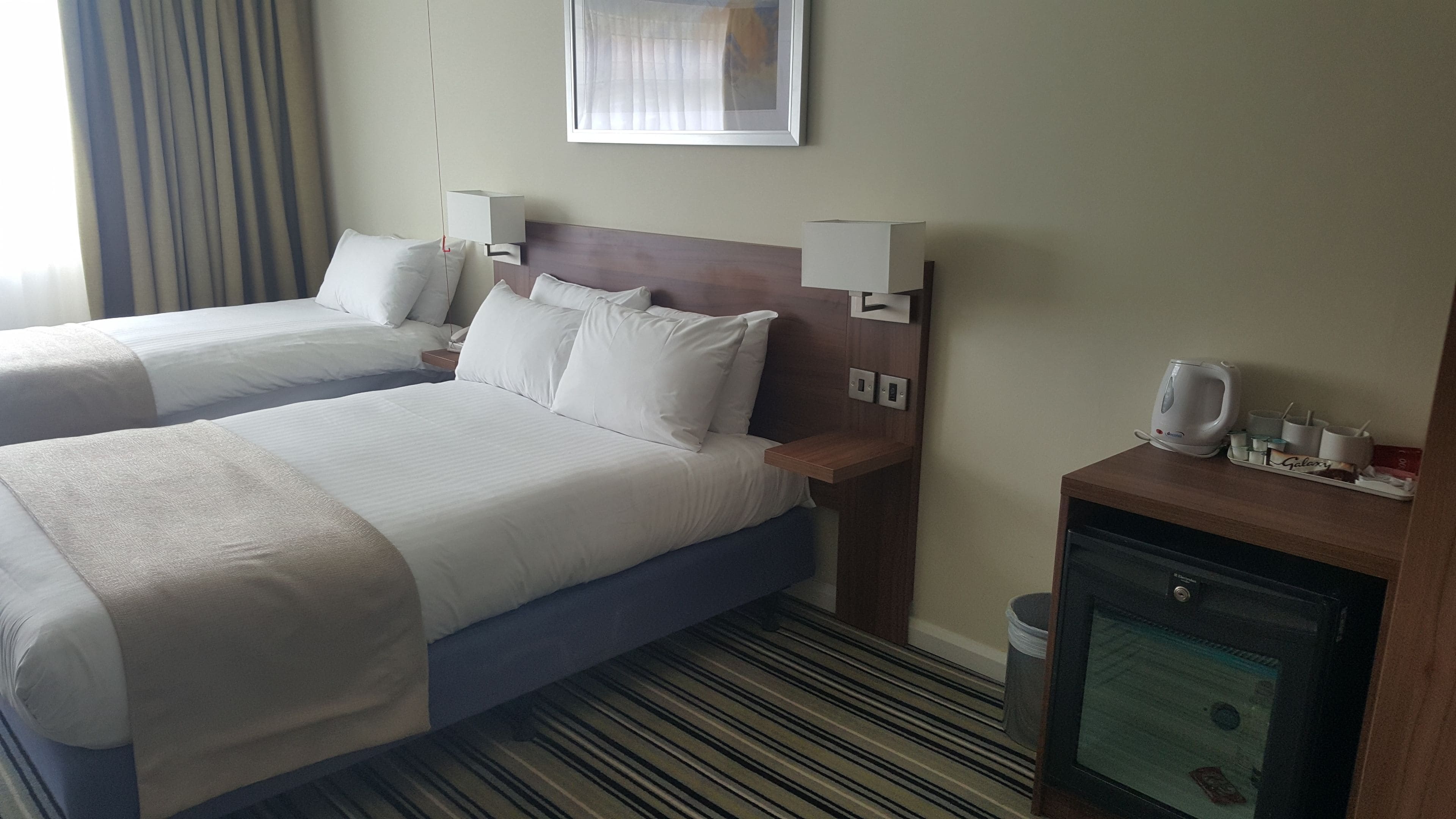 A hotel room with two beds, a nightstand, a small cabinet, and a window with curtains.
