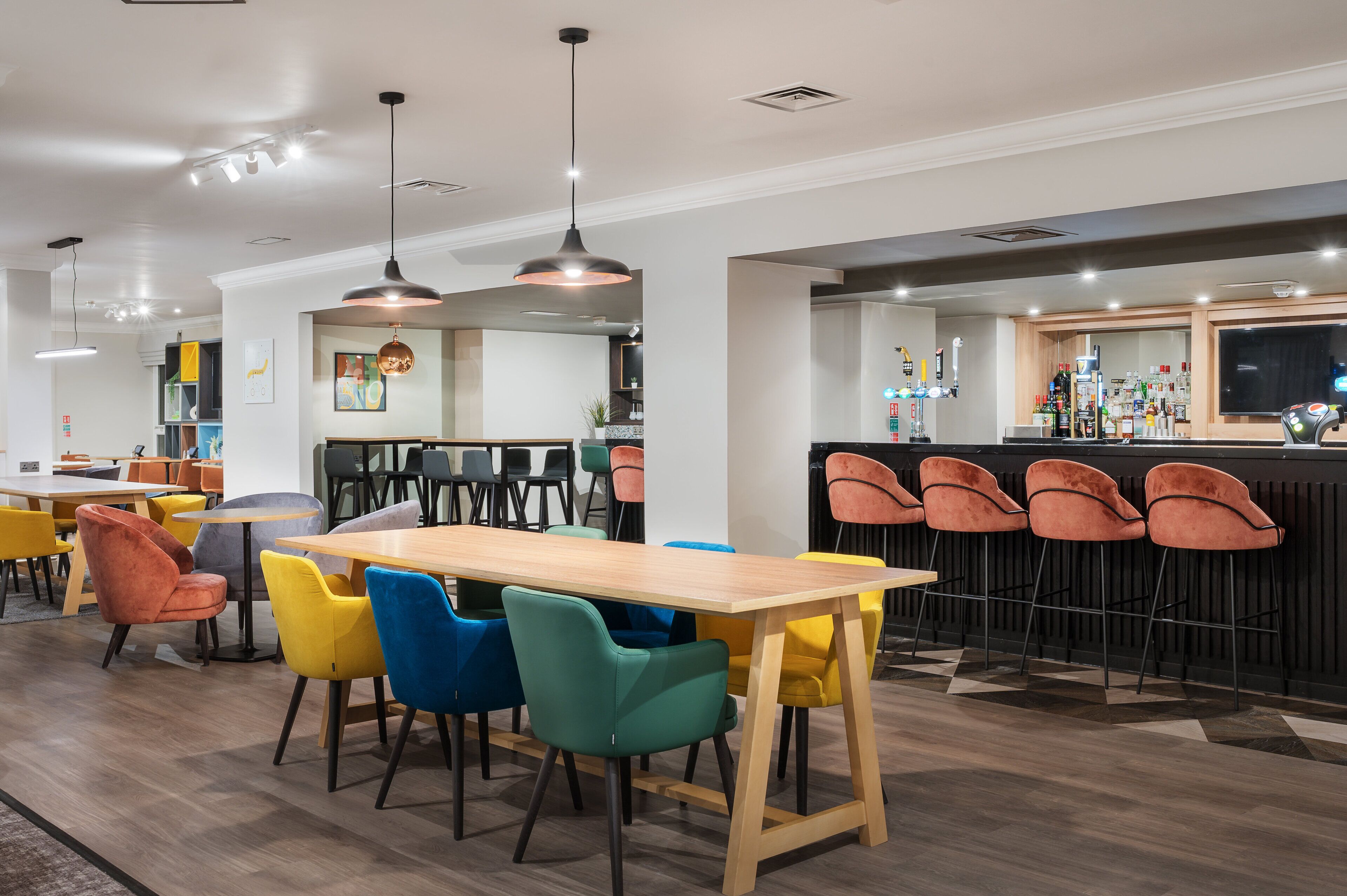 Un restaurante moderno con una mesa de madera y sillas coloridas, una zona de bar con taburetes e iluminación colgante.