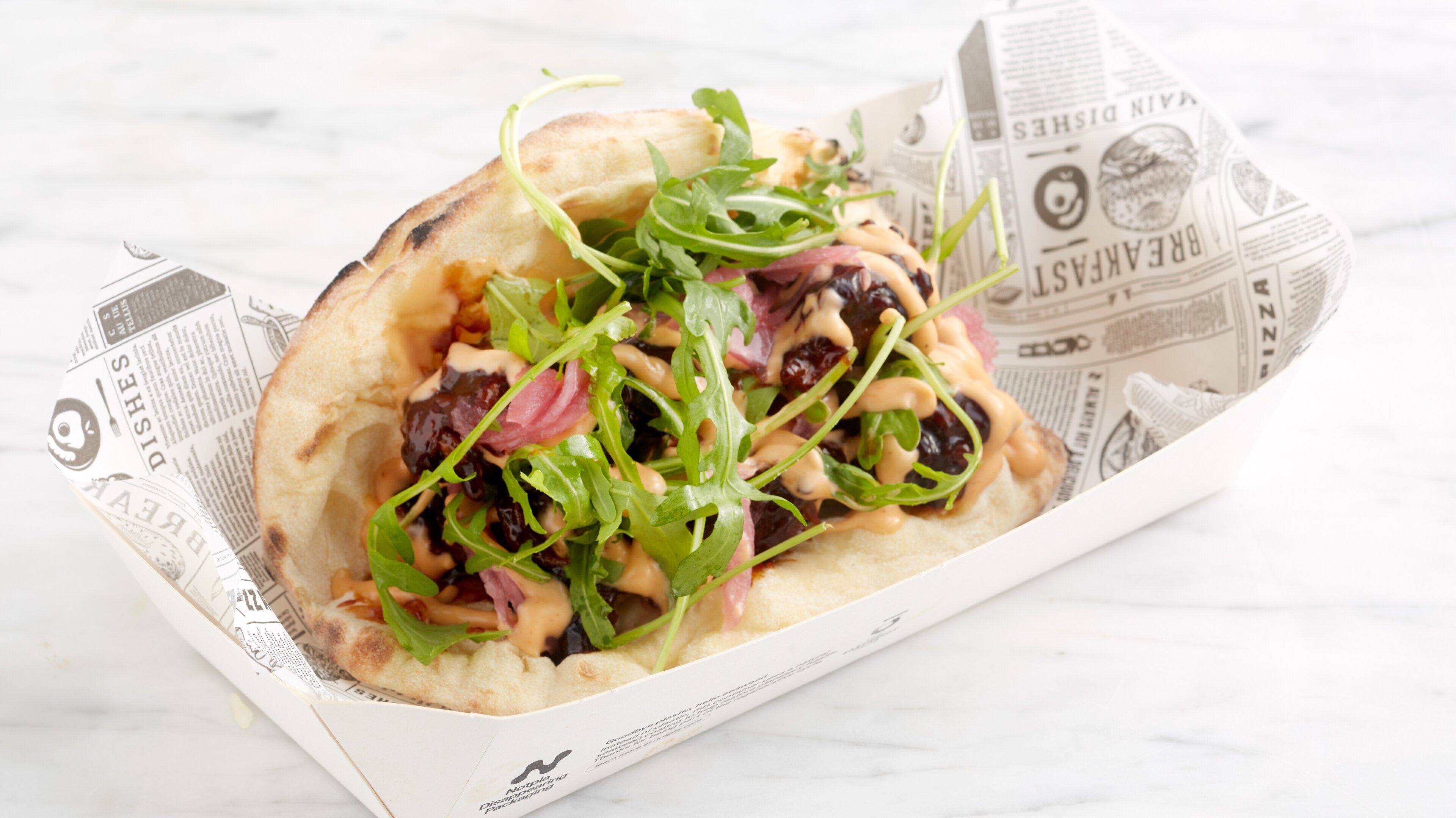 A flatbread sandwich with greens and a creamy sauce, served on a newspaper-lined tray.