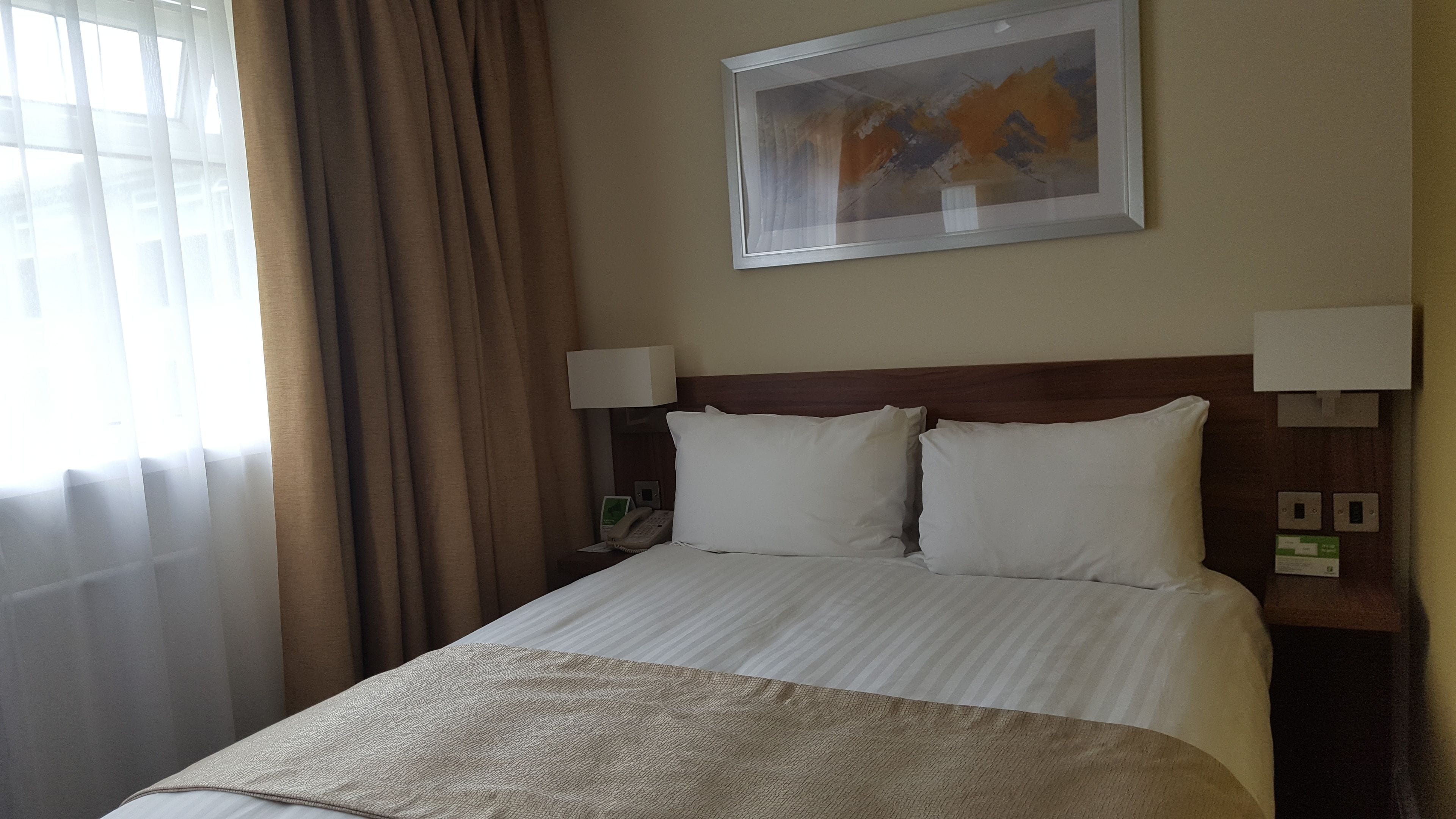 Una habitación de hotel con una cama, dos lámparas de noche, un cuadro en la pared y una ventana con cortinas.