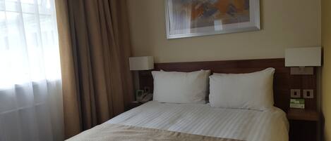 Una habitación de hotel con una cama, dos lámparas de noche, un cuadro en la pared y una ventana con cortinas.