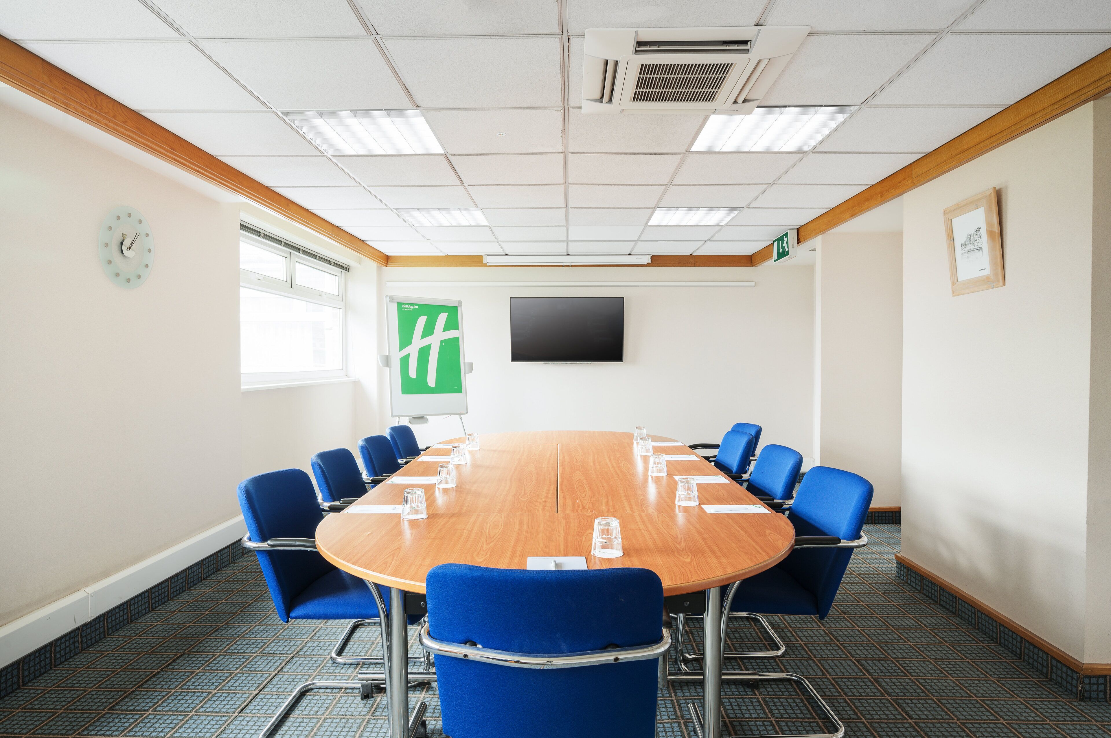A conference room with a wooden table, blue chairs, a clock, a TV, and a green exit sign.