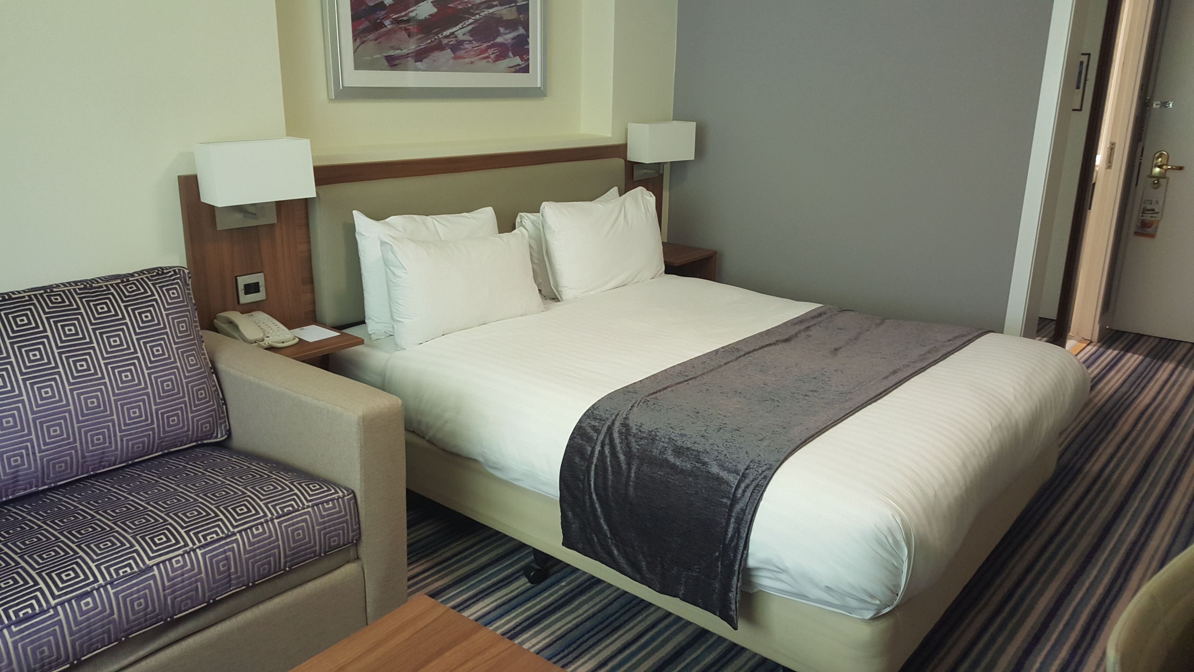 Una habitación de hotel con una cama, una mesita de noche con teléfono, una silla y una lámpara.