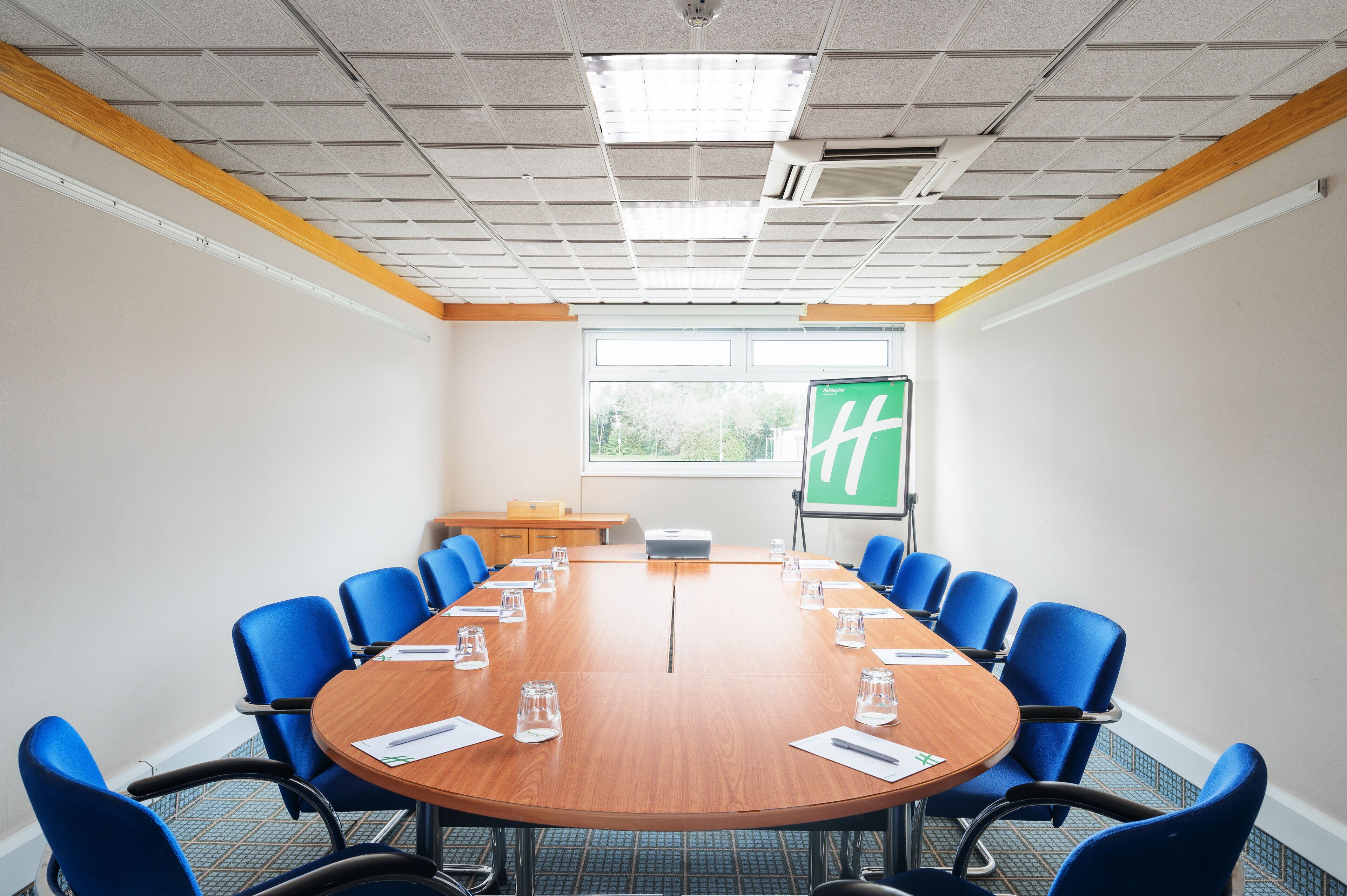 Un salón de conferencias con una mesa de madera, sillas azules, una pantalla de proyector y una ventana con vistas a zonas verdes.