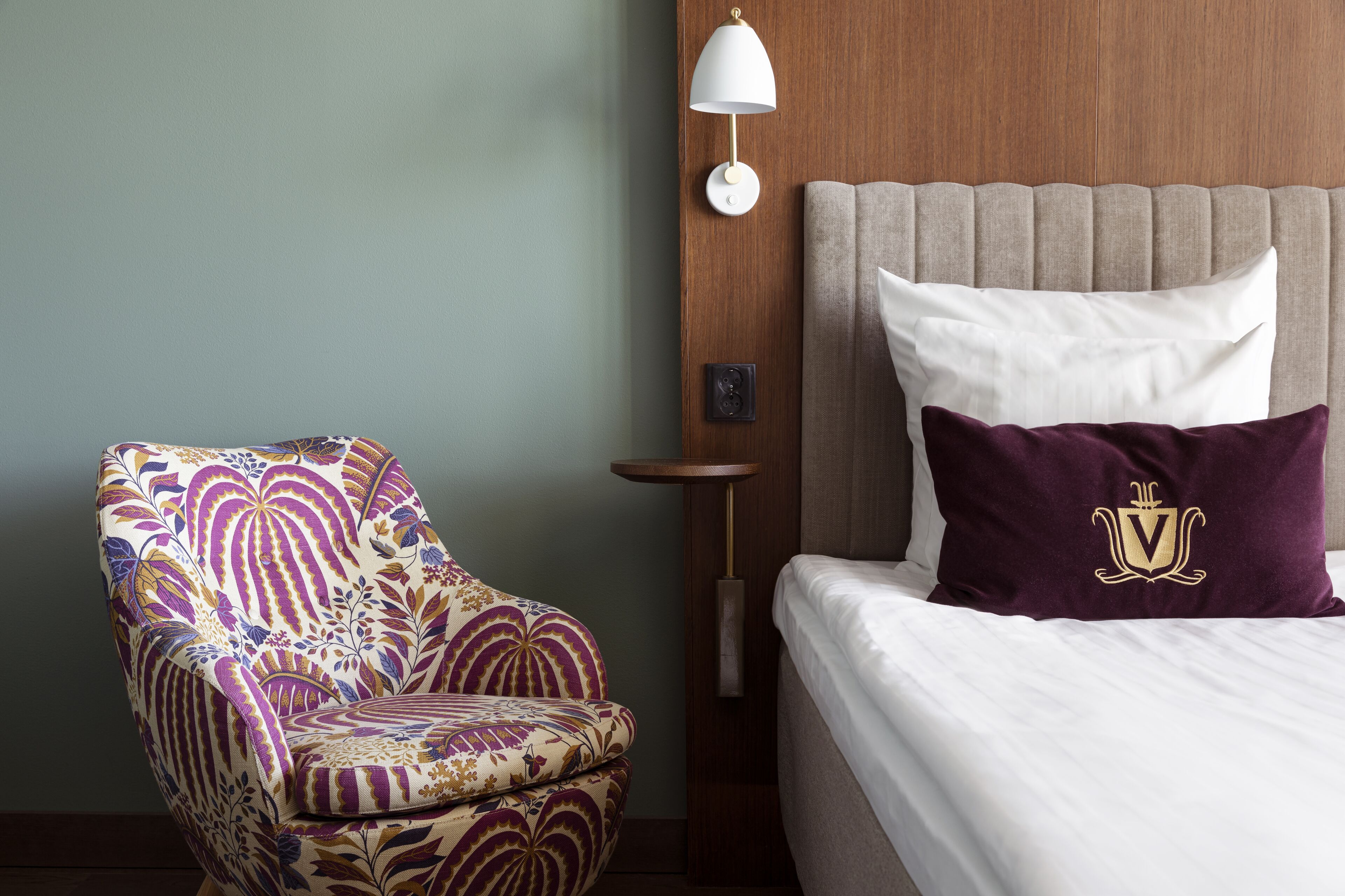 A hotel room with a patterned armchair, a bed with white and purple bedding, and a wall-mounted lamp.