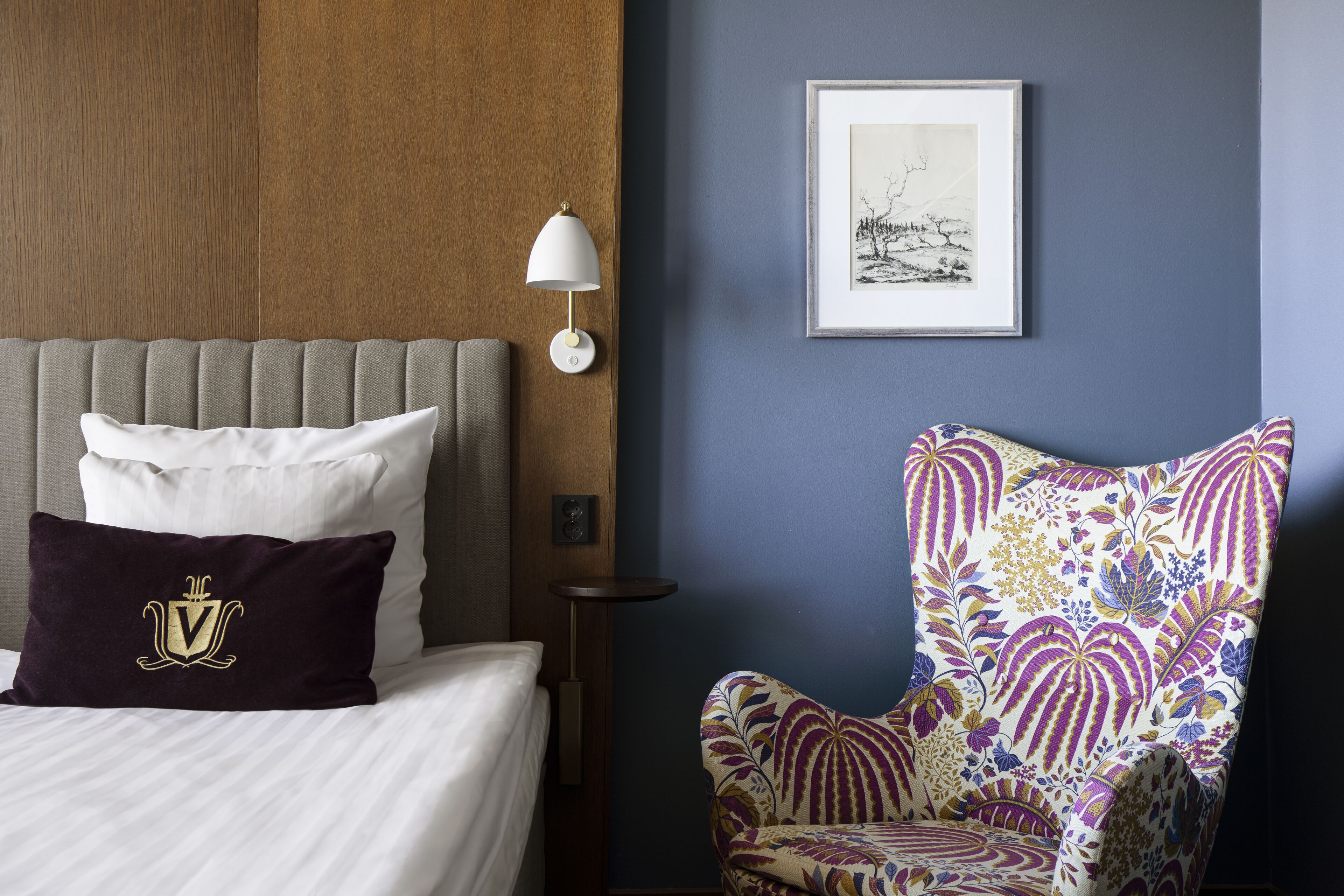 Una habitación de hotel con una cama, un sillón estampado, una lámpara de pared y un cuadro enmarcado.
