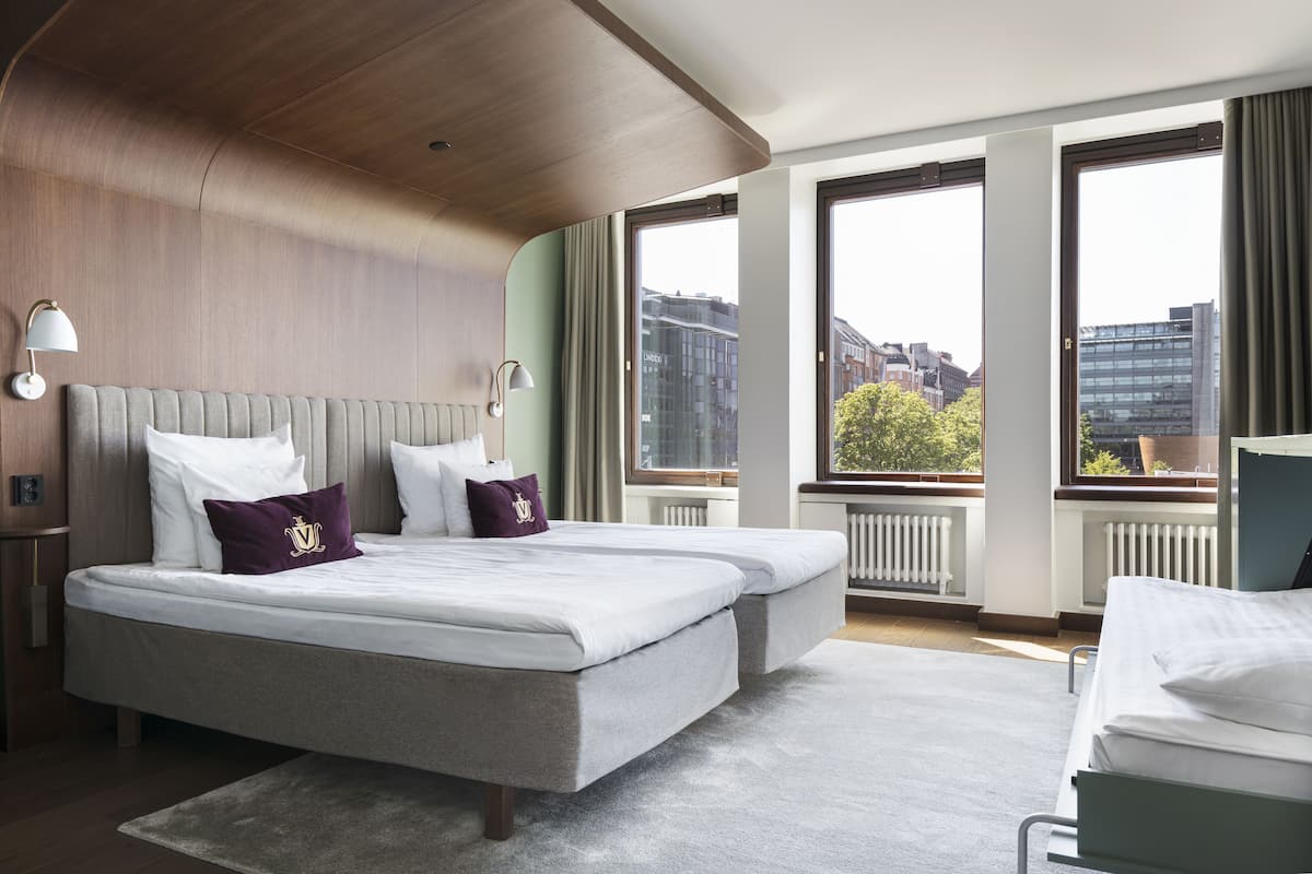 Habitación de hotel con una cama grande, dos mesitas de noche, un ventanal con vistas a la ciudad y techo de madera.