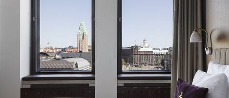 Habitación de hotel con cama, dos ventanas, una lámpara y vistas a edificios de la ciudad.