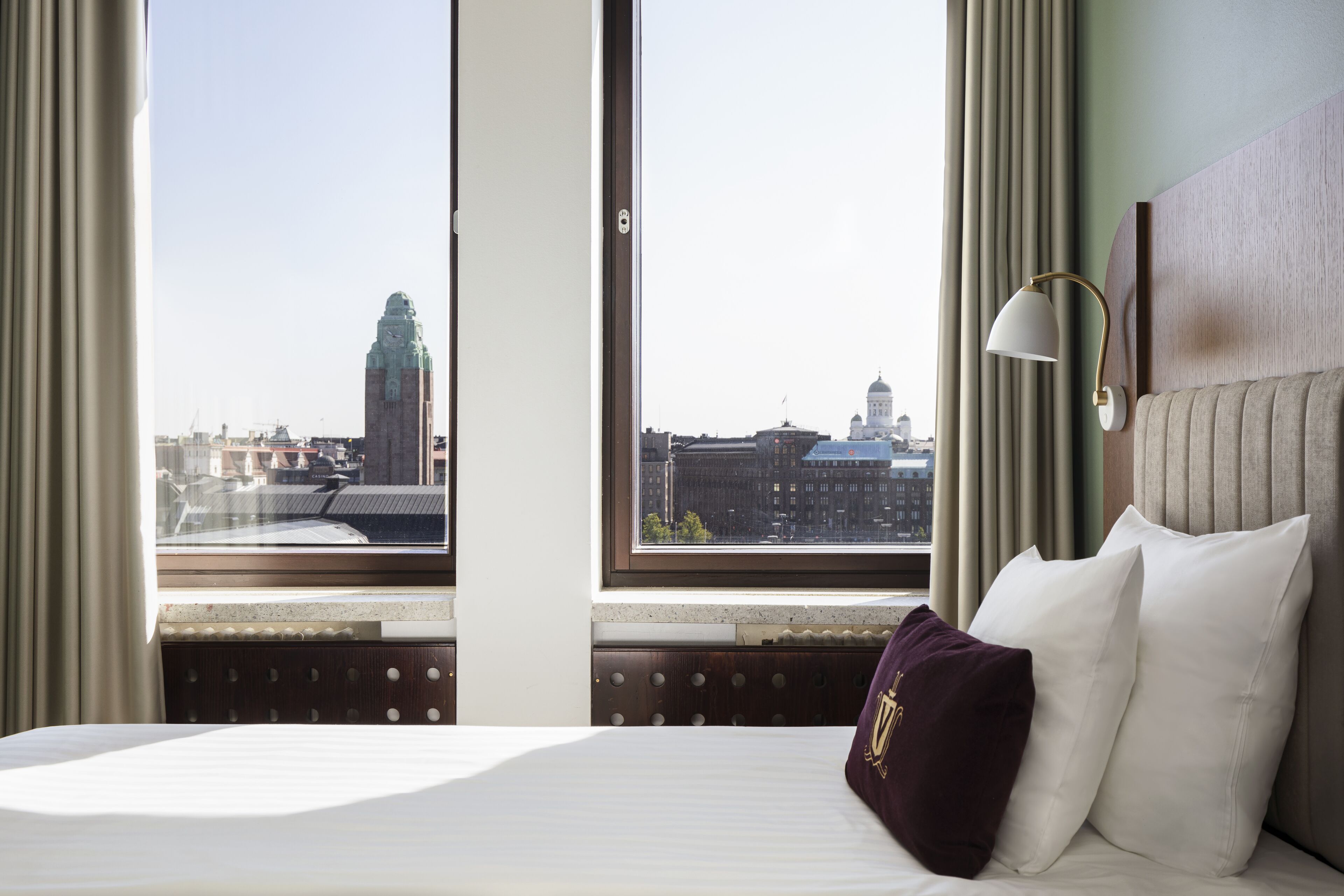 A hotel room with a large window, a bed with white linens, a headboard, and a lamp.