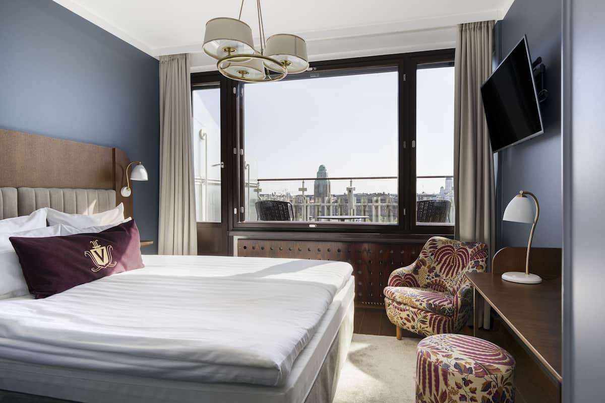 Habitación de hotel con una cama grande, un escritorio, una silla, un televisor y vistas a la ciudad a través de un ventanal amplio.