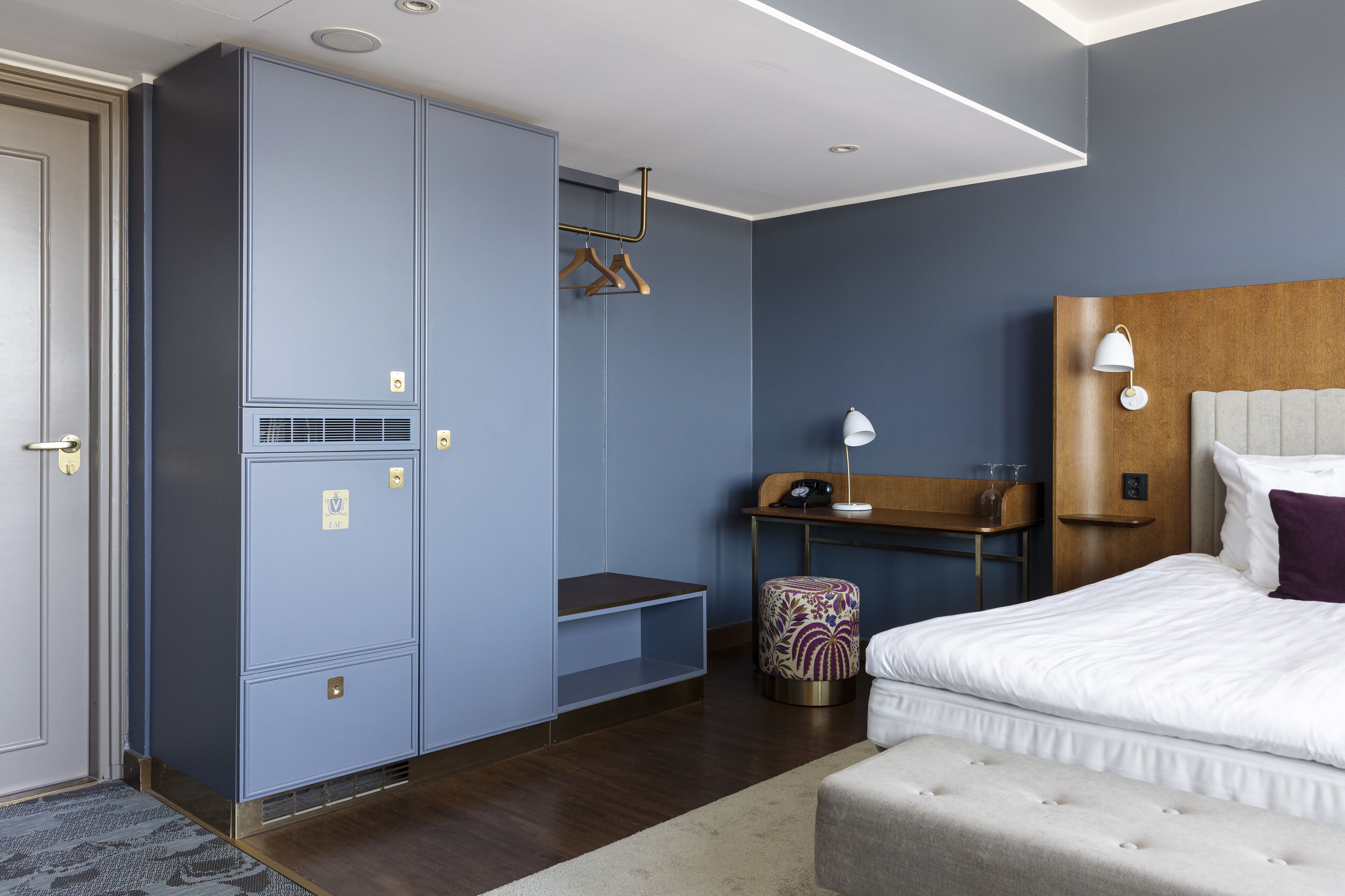 A modern hotel room with a large wardrobe, a desk with a lamp, a bed with a headboard, and a small stool.
