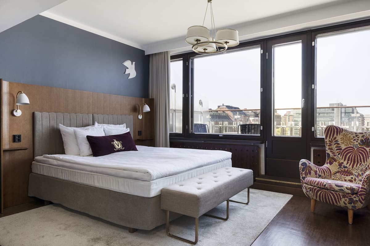Habitación de hotel moderna con una cama grande, un banco, un sillón estampado y vistas a la ciudad a través de amplias ventanas.