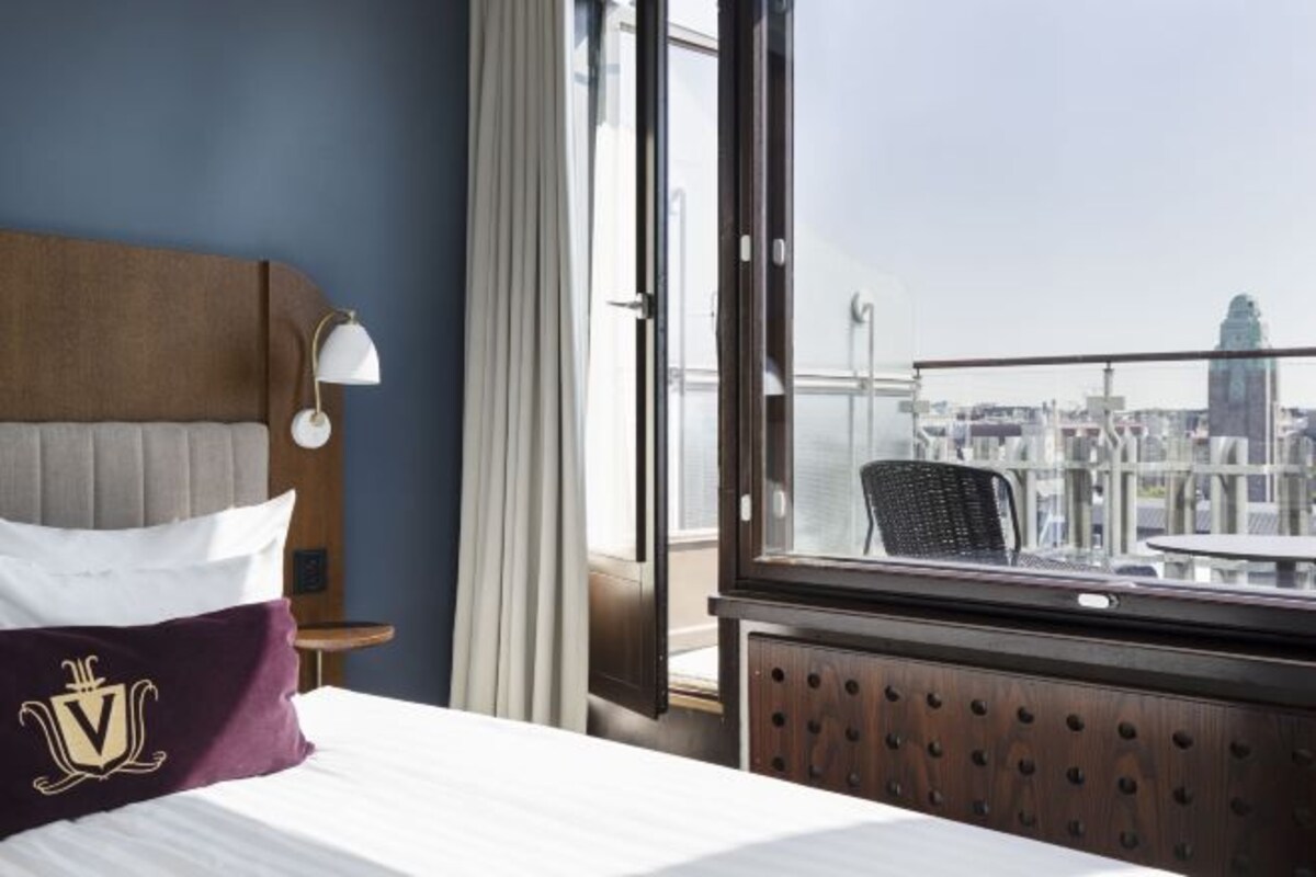 Habitación de hotel con cama, mesita de noche, lámpara, ventana con vista a la ciudad y balcón con silla y mesa.