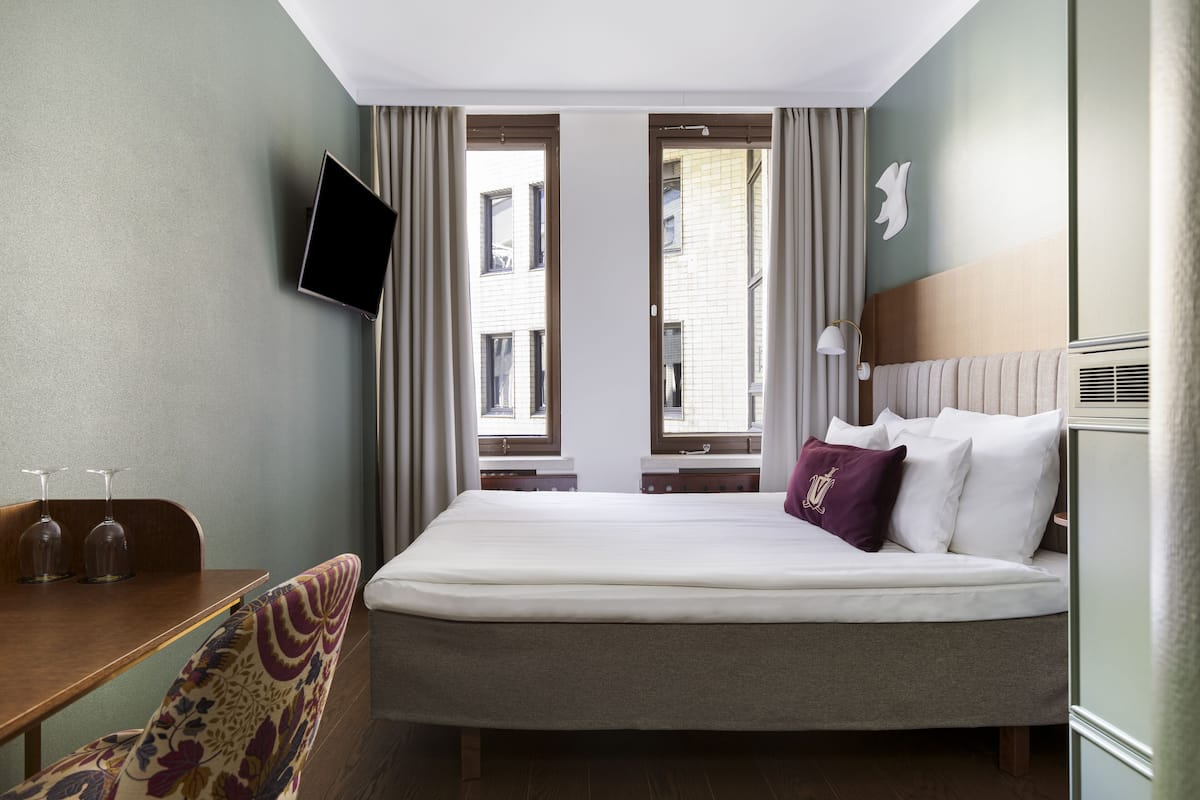 Una habitación de hotel con una cama, un escritorio con silla, un televisor montado en la pared y una ventana con cortinas.