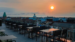 Terraza en la azotea con mesas y sillas, con vistas a un paisaje urbano de edificios con cúpulas y un atardecer.