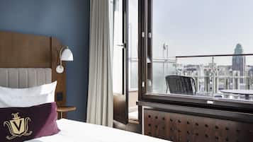 Quarto de hotel com cama, mesa de cabeceira, abajur, janela com vista para a cidade e uma cadeira perto da janela.