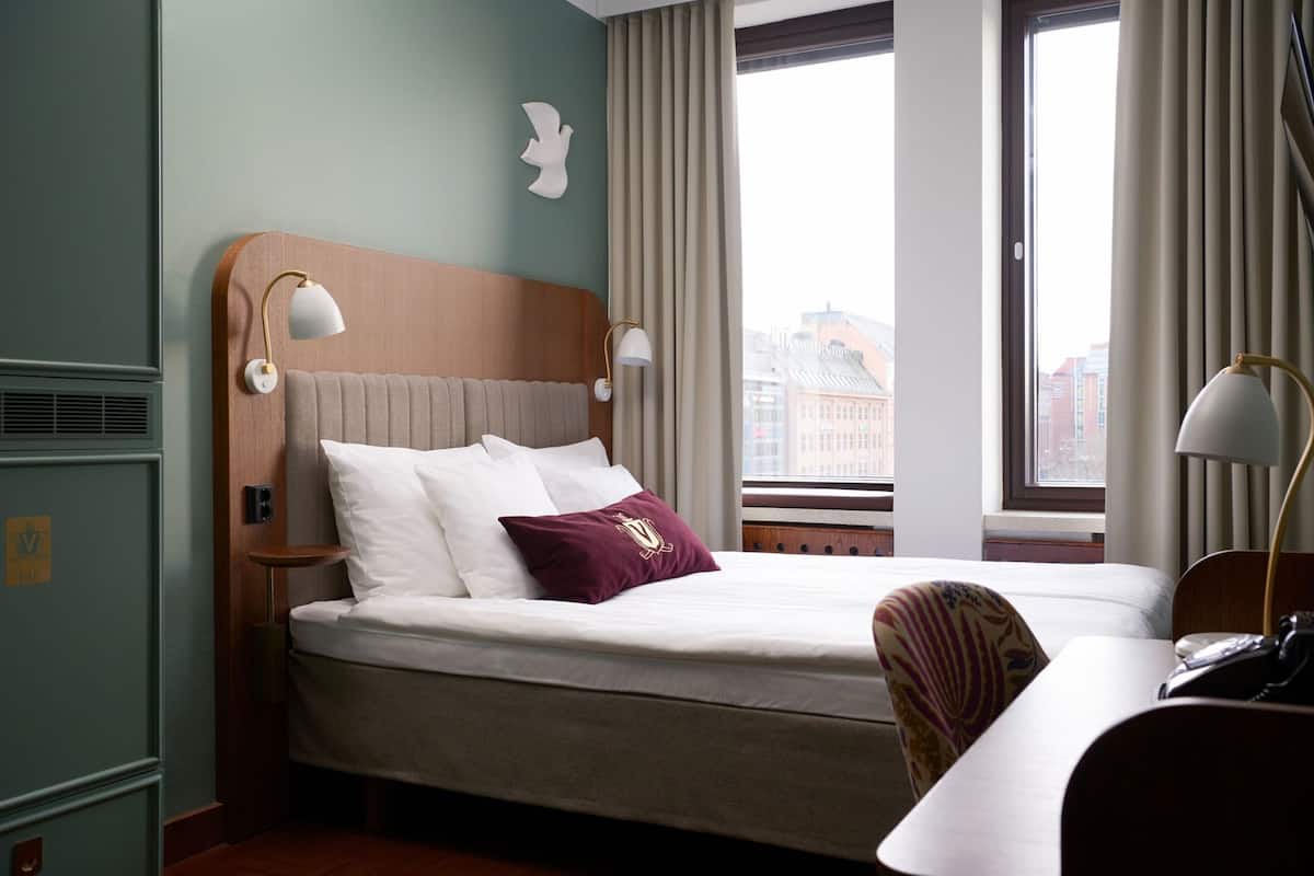 Habitación de hotel con cama, dos lámparas de noche, una silla, escritorio, ventana con cortinas y vistas a la ciudad.