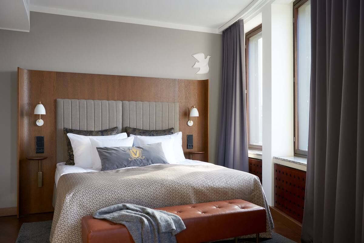 Habitación de hotel con una cama grande, cabecero de madera, dos lámparas de noche, un banco y una ventana con cortinas.