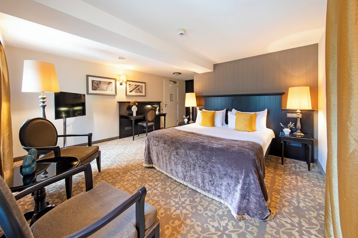 Ein Hotelzimmer mit einem großen Bett, zwei Nachttischen mit Lampen, einem Schreibtisch mit Stuhl, einem an der Wand befestigten Fernseher und einem gemusterten Teppich.