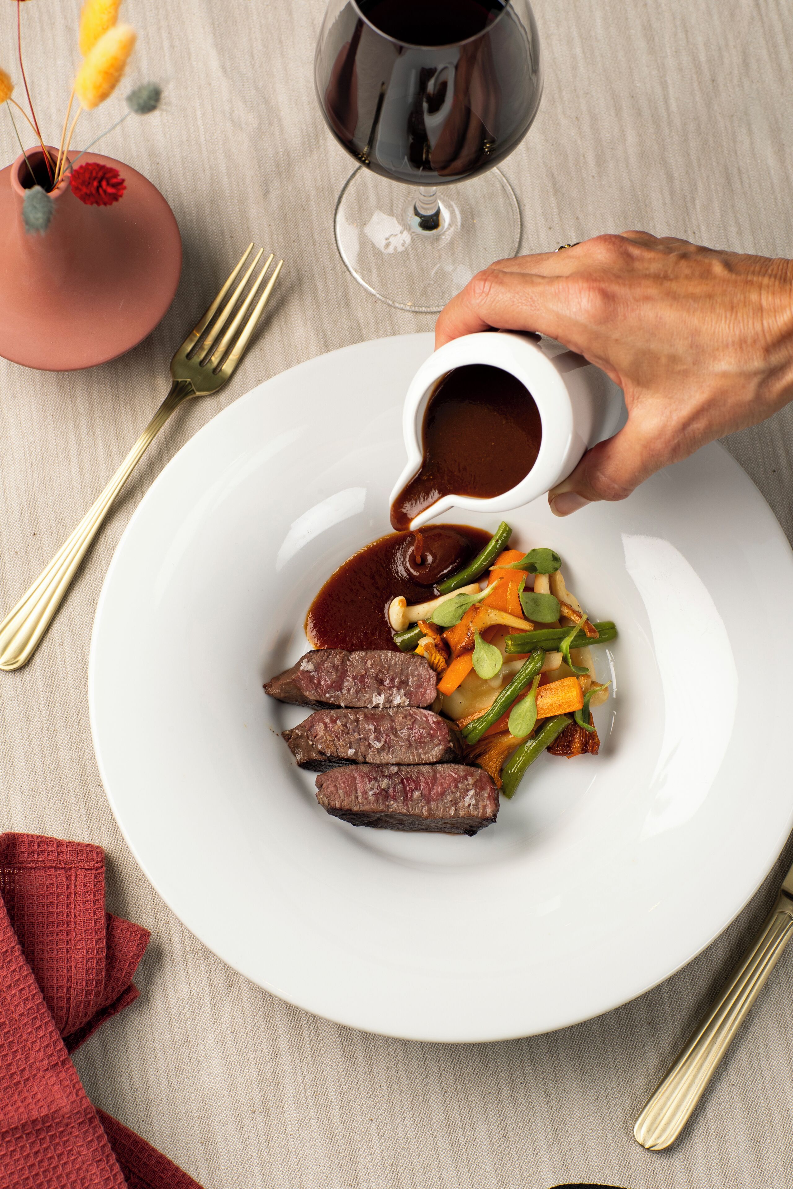 A white plate with sliced meat, vegetables, and a sauce being poured.