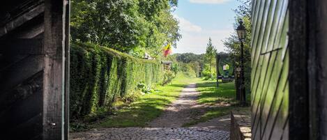 A cobblestone path leading through a lush green area with a flag on a pole, a wooden structure on the left, and a metal gate on the right.