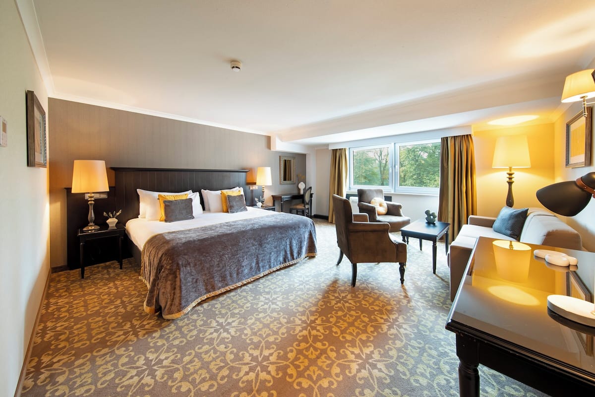 Ein Hotelzimmer mit einem großen Bett, einem Schreibtisch, einem Sessel und einem kleinen Tisch. Ein Fenster mit Vorhängen bietet Blick auf Bäume.