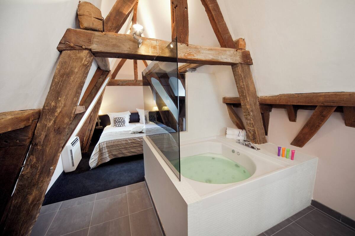 Ein Schlafzimmer mit großem Fenster, Holzbalkendecke und einer Badewanne mit Glastrennwand.