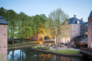 A riverside courtyard with outdoor seating, a brick building, and a bridge. - Bilderberg Chateau Holtmuhle (Tegelen)