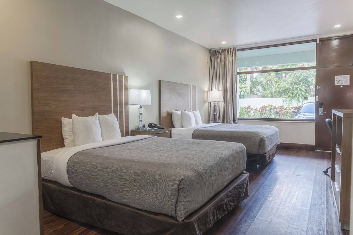 A hotel room with two beds, a desk, a chair, a lamp, and a large window with curtains.