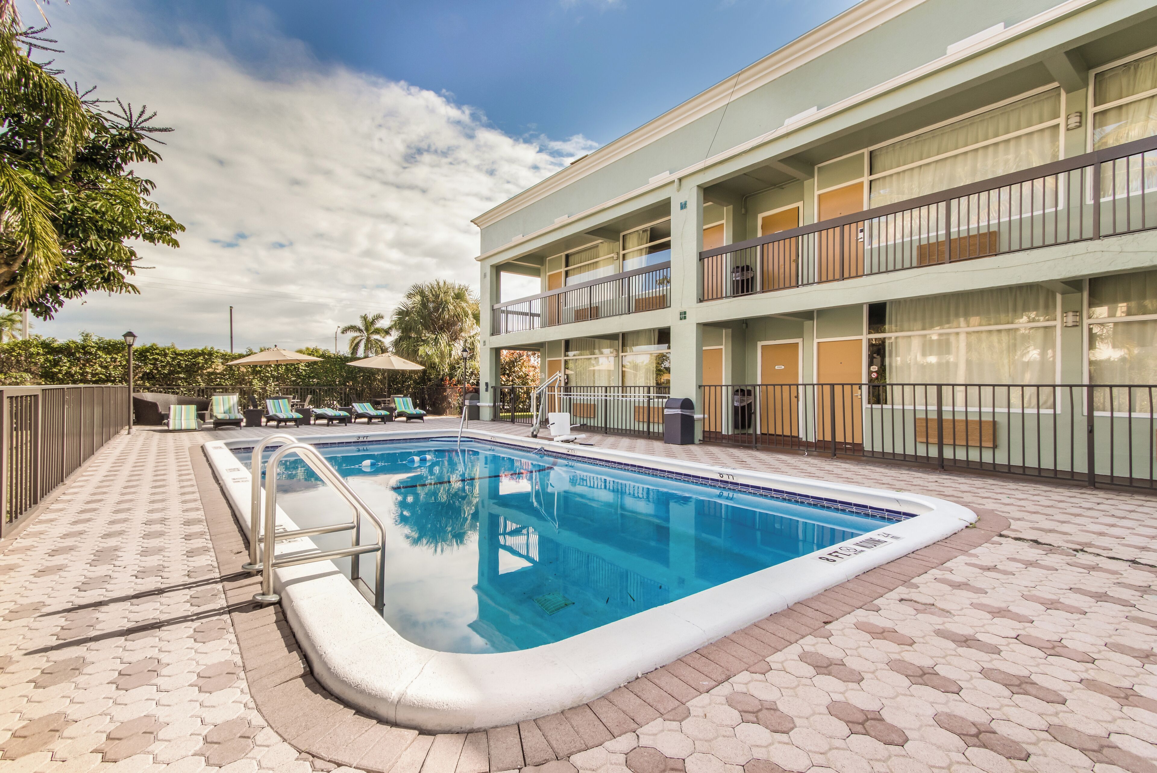 Una piscina con plataforma de azulejos, un edificio de varios pisos con balcones y una palmera al fondo.