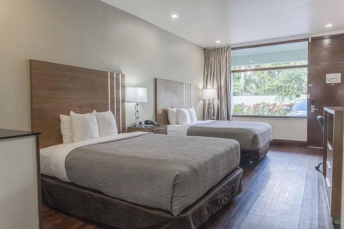 A hotel room with two beds, a desk, a chair, a window with curtains, and a view of greenery outside.