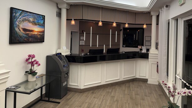 A reception area with a modern design, featuring a reception desk, an ATM, a large framed artwork, and a decorative plant. - Boca By the Sea (Boca Raton)