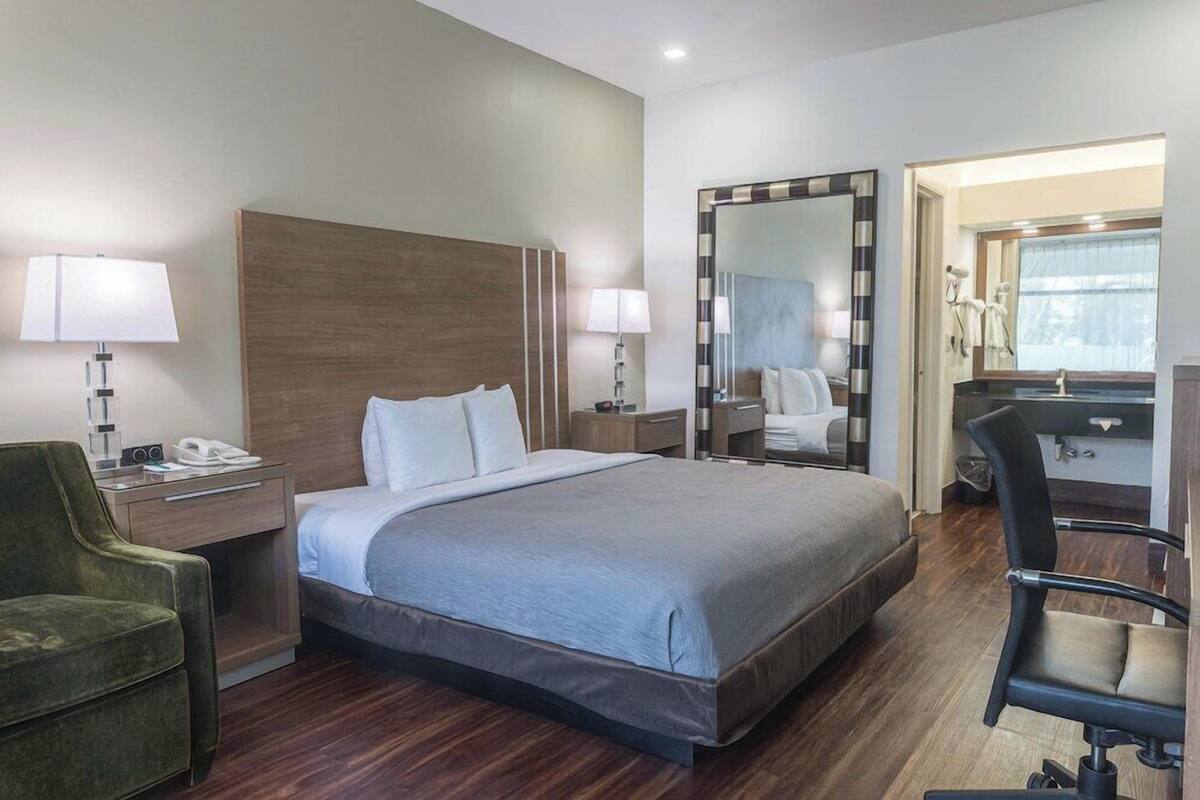 A hotel room with a large bed, bedside tables, a green armchair, a mirror, and a desk with a chair.