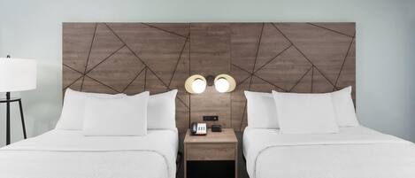 A hotel room with two beds, a wooden headboard, bedside lamps, a nightstand with a phone and a remote control, and a lamp on a side table.