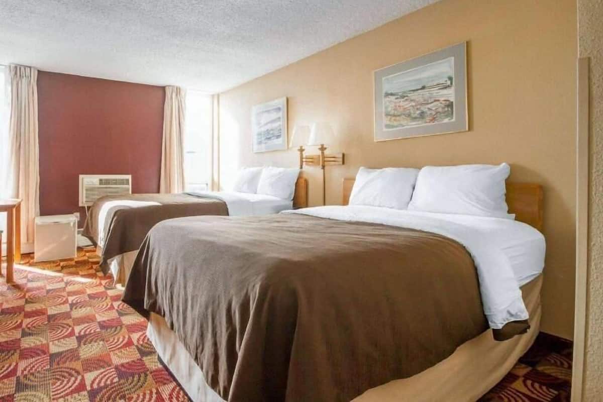 A hotel room with two beds, a desk, a chair, a lamp, and a painting on the wall.