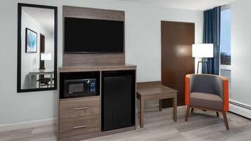 Ein Hotelzimmer mit Flachbildfernseher, Minikühlschrank, Mikrowelle, einem Holztisch, einem Sessel und einem großen Fenster mit Vorhängen.